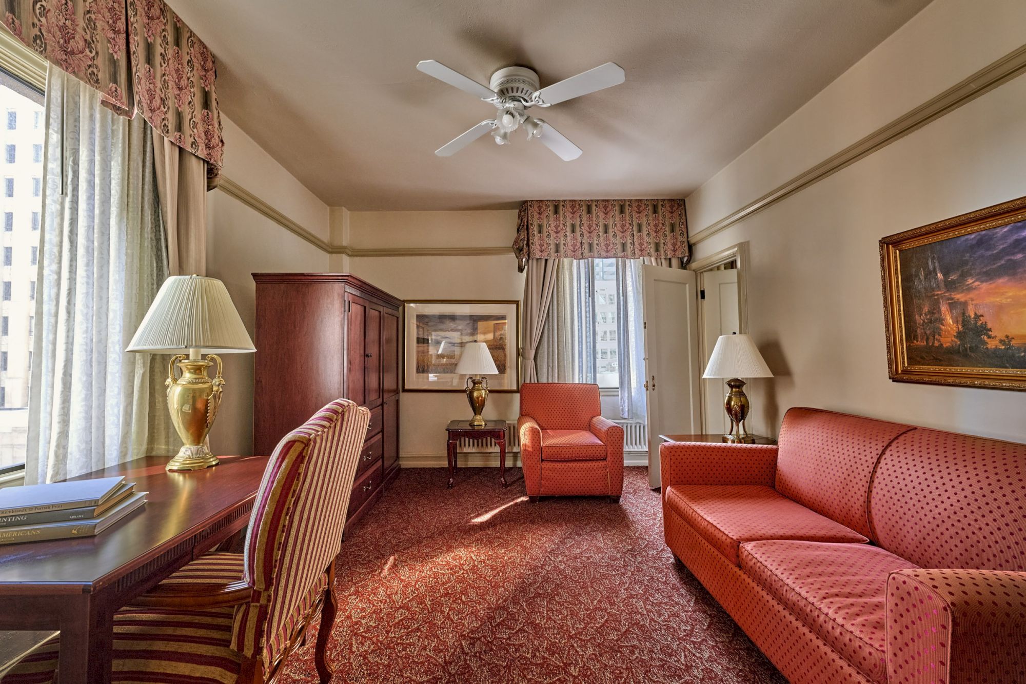 A cozy hotel room with a desk, chairs, sofa, and lamps. Ceiling fan overhead; framed artwork and large windows with curtains.