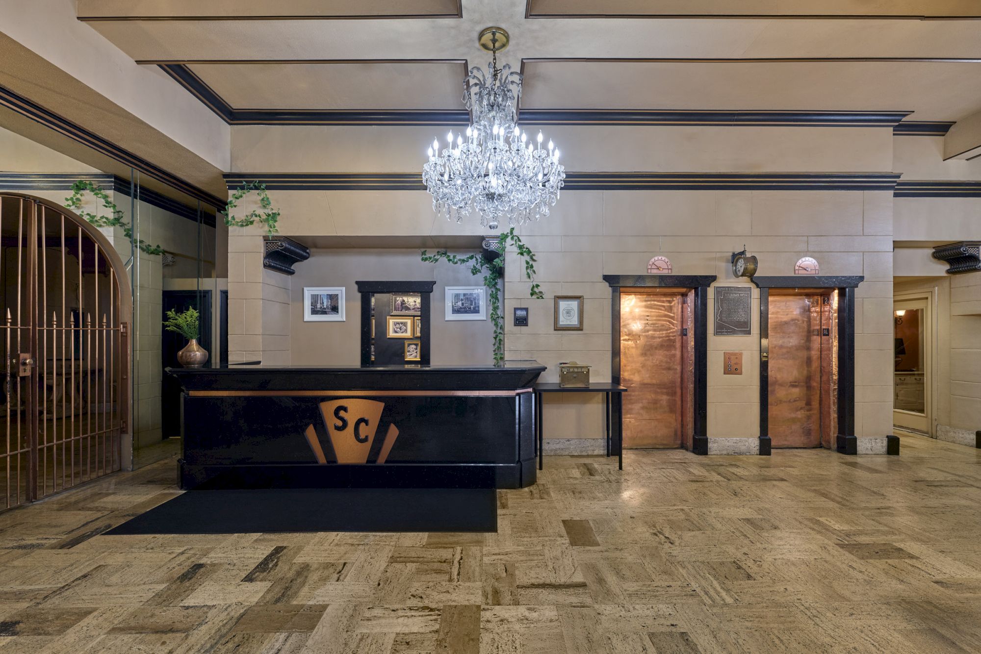 A lobby with a chandelier, reception desk, two elevators, and framed pictures on the walls, evoking an elegant and classic ambiance.