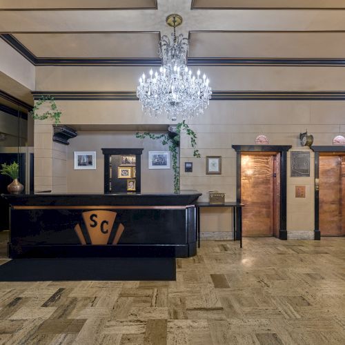 The image shows a lobby with a chandelier, a reception desk, two elevators, and framed pictures on the walls.