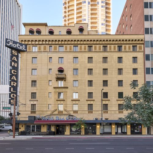 The image shows a historic hotel building with a "Hotel San Carlos" sign, surrounded by modern high-rise buildings and a tree-lined street.