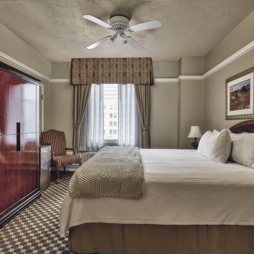 A cozy hotel room with a neatly made bed, large wardrobe, two side tables, armchair, curtain-covered window, ceiling fan, and framed art.