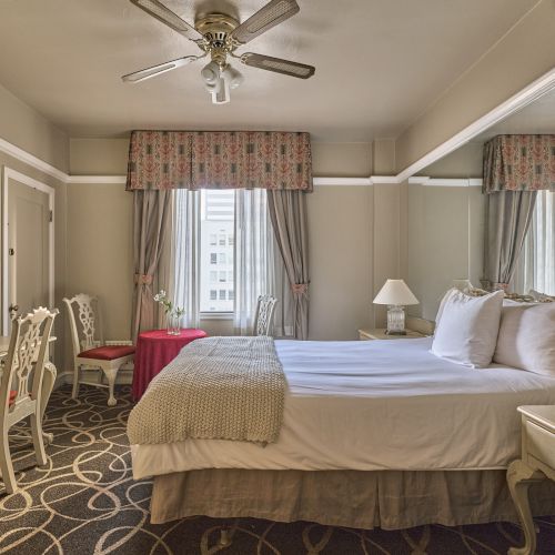 A well-decorated bedroom with a bed, bedside tables, mirrors, and a desk. The room features neutral tones, patterned carpet, and a ceiling fan.