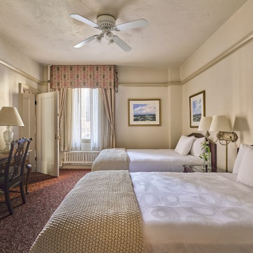 A hotel room with two beds, artwork on walls, a desk, chair, and lamps. Ceiling fan and window with drapes complete the cozy ambiance.