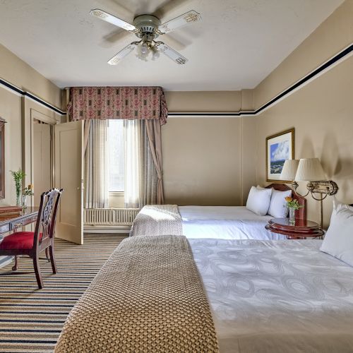 The image shows a well-lit hotel room with two beds, a desk with a chair, artworks on the walls, and a ceiling fan.