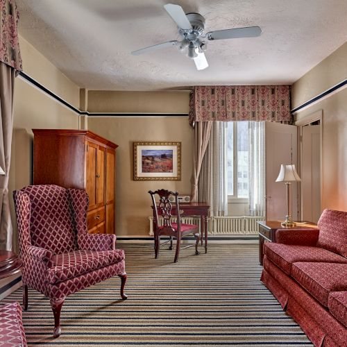 The image shows a living room with patterned furniture, striped carpet, a ceiling fan, and framed artworks on the walls.