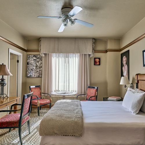 A cozy hotel room with a bed, two chairs, a desk, lamps, and wall art. A ceiling fan and window provide light and ventilation.