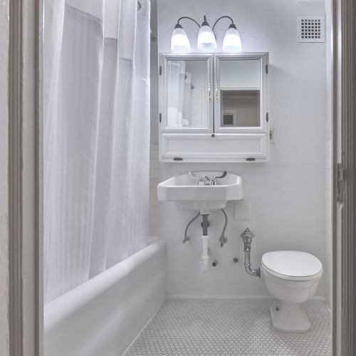 A small, white bathroom with a sink, toilet, and a shower-tub combo. There’s a mirrored cabinet and overhead lights.