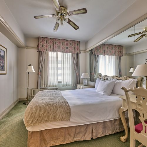 A cozy bedroom with a neatly made bed, mirrored wall, ceiling fan, and vintage decor, featuring lamps and floral curtains.