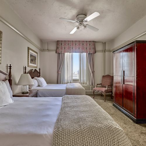 The image shows a hotel room with two beds, a ceiling fan, a wardrobe, and a window with curtains, offering a cozy and classic look.