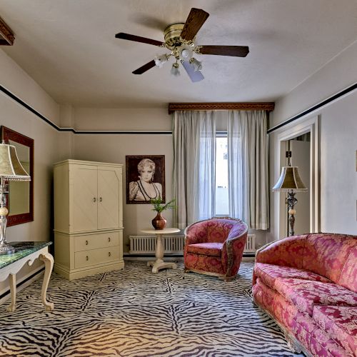 A vintage-style room with patterned sofas, zebra-print rug, and art on the walls. A ceiling fan, lamps, and curtains complete the decor.