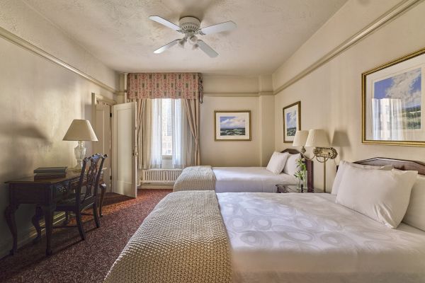 The image shows a hotel room with two beds, a desk, lamps, and framed art on the walls, featuring beige and neutral tones.