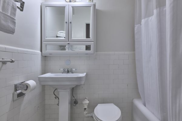 A small, clean bathroom with a pedestal sink, toilet, mirrored cabinet, and a bathtub with a shower curtain. White tiles cover the floor and walls.