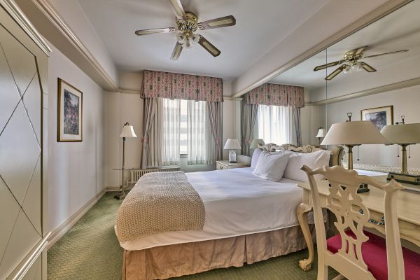 A well-decorated bedroom with a double bed, mirrored wall, ceiling fan, and lamps, featuring floral curtains and carpeted flooring.