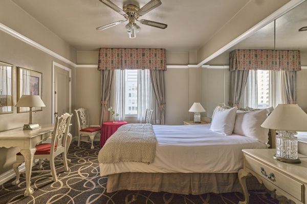 The image shows a cozy bedroom with a double bed, elegant furniture, a ceiling fan, and a window with curtains. The decor is classic and inviting.