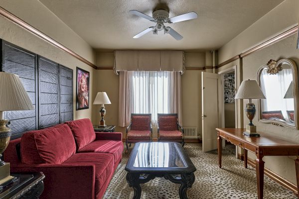 A cozy living room with red furniture, a glass coffee table, decorative lamps, a ceiling fan, and art pieces on the walls.