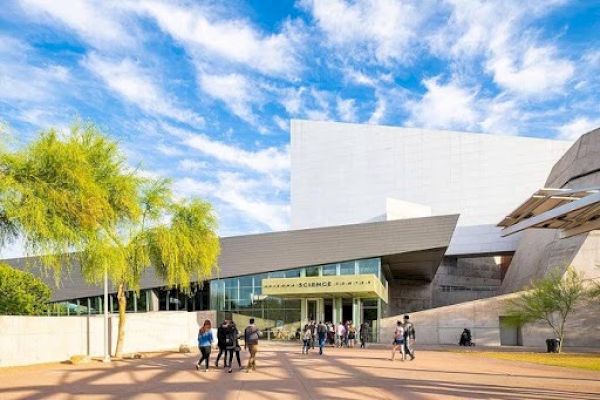 The image shows a modern building complex with people walking outside under a bright blue sky with clouds.