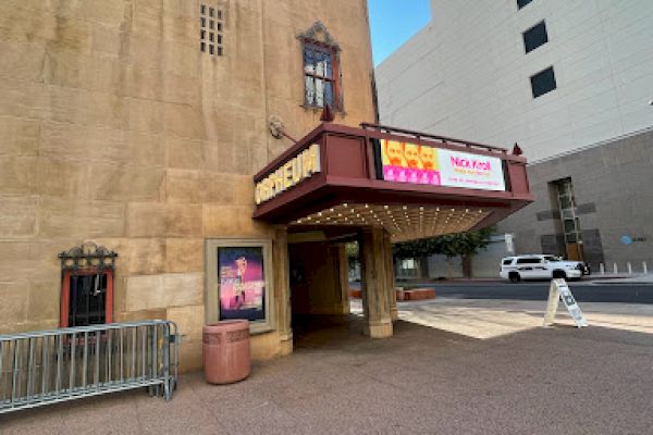 A vintage theater entrance with ornate architecture and a marquee sign. Posters are displayed, and nearby buildings are visible.