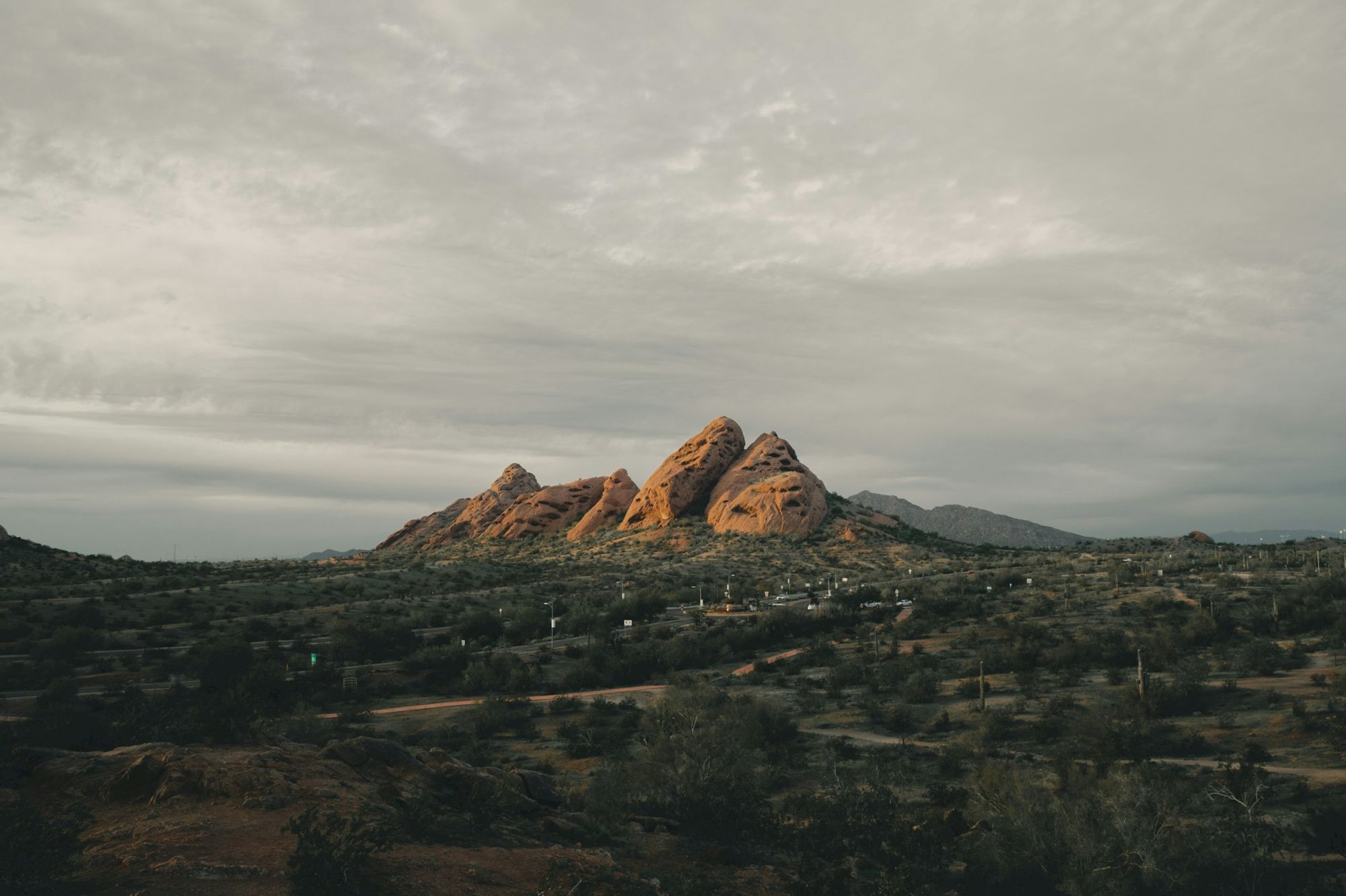 A desert landscape with rugged mountains under a cloudy sky. The terrain is rocky and sparse, featuring some scattered vegetation.