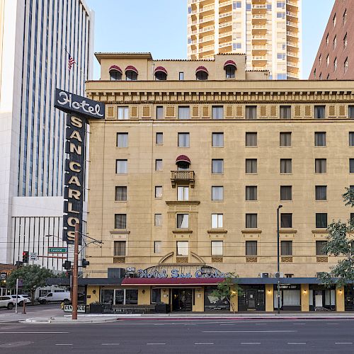 The image shows the Hotel San Carlos building, featuring a neon sign, surrounded by modern skyscrapers in an urban setting.