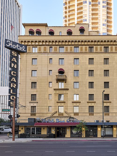 The image shows the Hotel San Carlos building, featuring a neon sign, surrounded by modern skyscrapers in an urban setting.