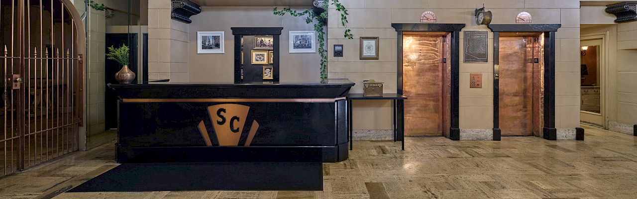 An elegant lobby with a chandelier, a reception desk, and two elevators. The floor is tiled, and framed pictures adorn the walls.