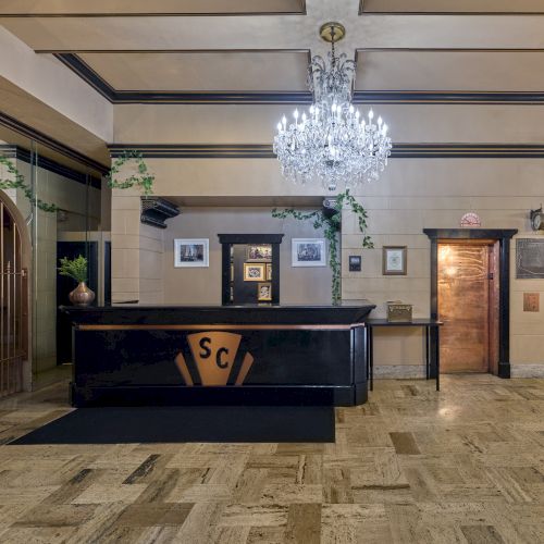 An elegant lobby with a chandelier, a reception desk, and two elevators. The floor is tiled, and framed pictures adorn the walls.