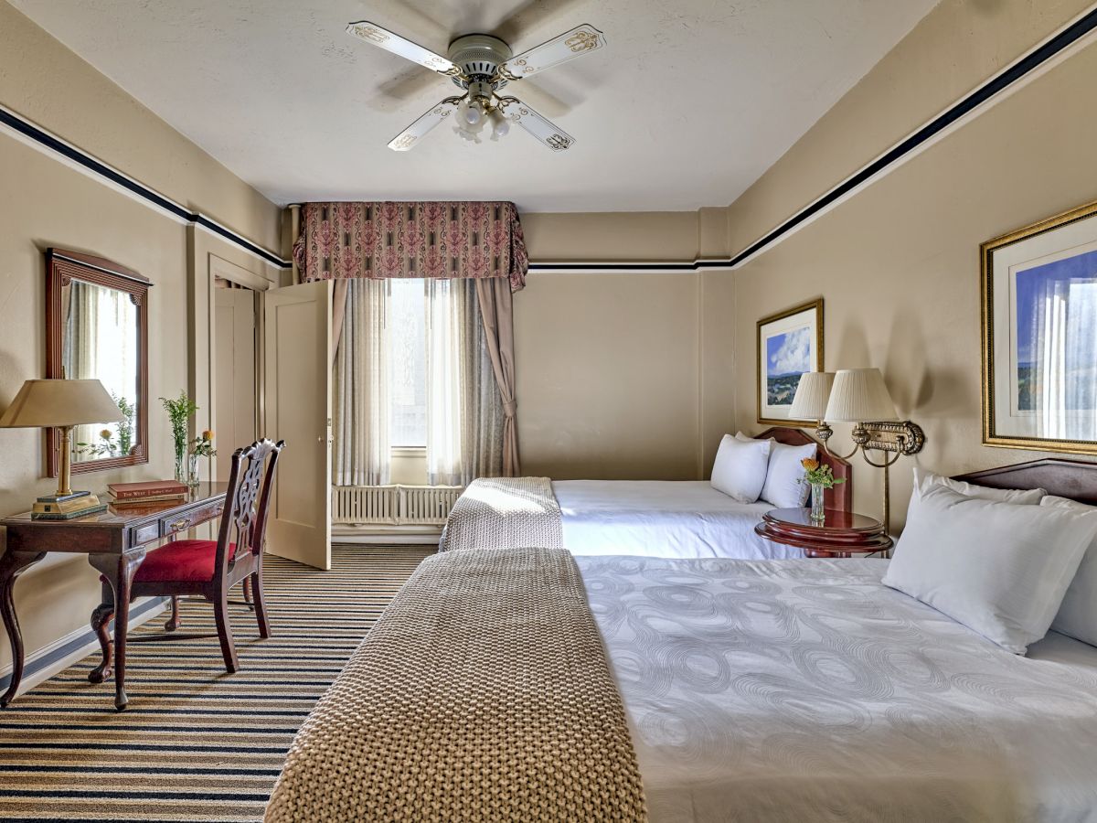 The image shows a hotel room with two beds, a desk, a chair, and wall art. There's a ceiling fan and a window with curtains.