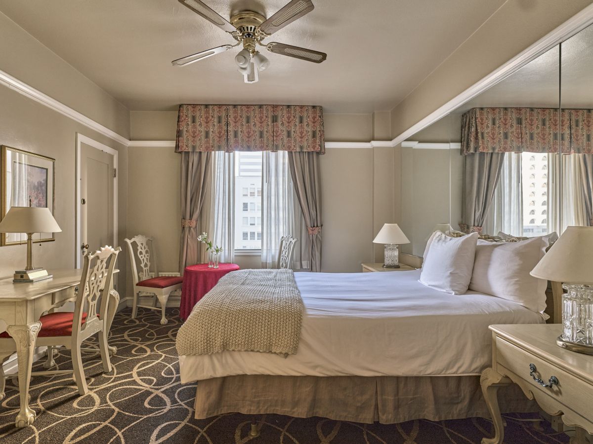 A well-decorated bedroom with a large bed, elegant furniture, lamps on side tables, patterned carpet, and a ceiling fan.