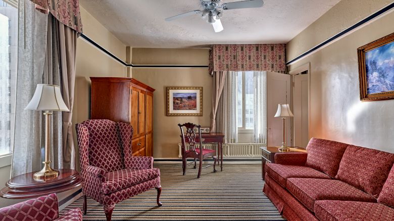 A cozy room features patterned chairs, a sofa, a wooden cabinet, a small table, and striped carpet, with paintings and curtains. Ceiling fan above.