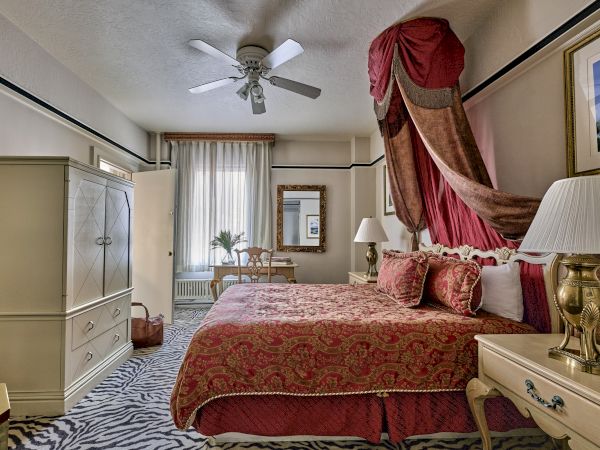 Elegant bedroom with a large bed, zebra-patterned carpet, ornate canopy, dresser, armchair, and ceiling fan, with natural light filtering in.