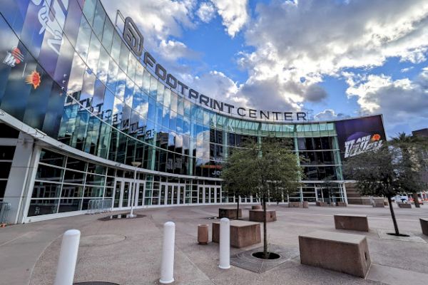 The image shows the exterior of the Footprint Center, a large building with glass windows and a clear blue sky with clouds above it.